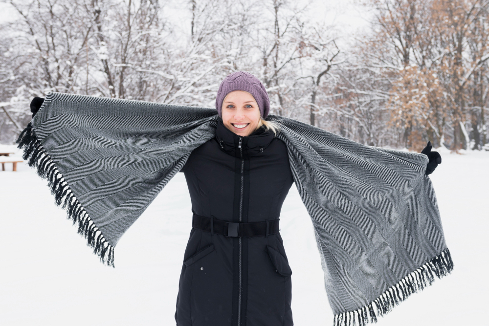 A person wearing a black winter coat and a purple hat stands in a snowy landscape, holding a large gray scarf out to the sides.