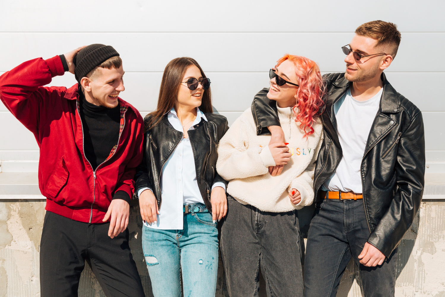 A group of four stylish young adults dressed in jackets and casual wear, posing in bright sunlight.