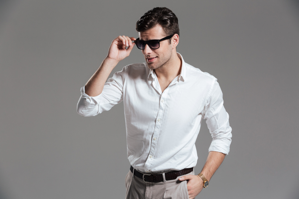 A well-dressed man in a white button-up shirt and beige pants poses confidently, adjusting his sunglasses .