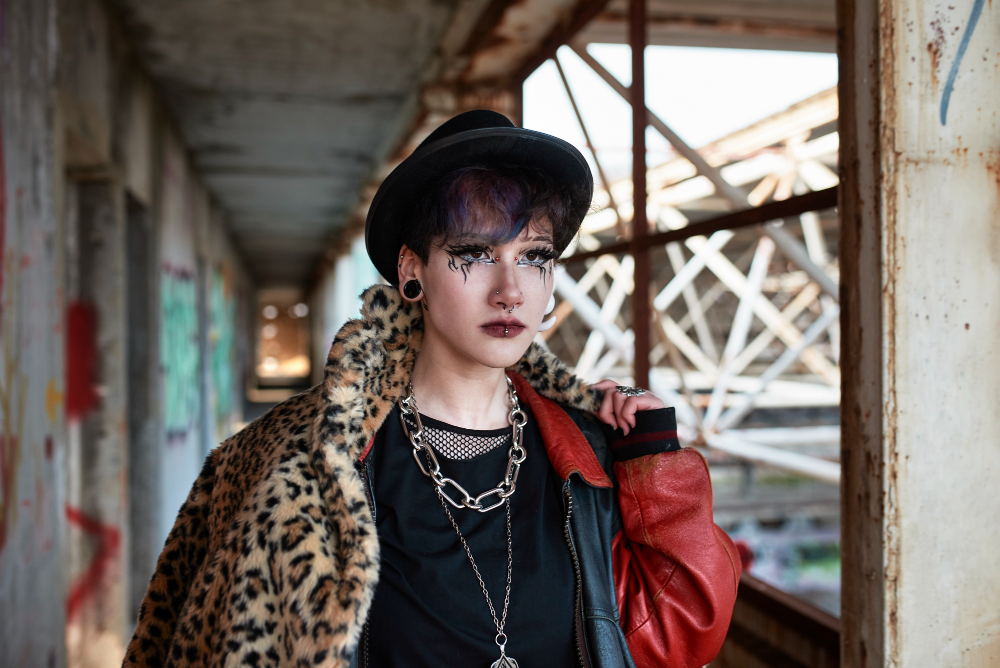 A person wearing a leopard print coat, red leather jacket, and a black hat stands in an industrial corridor with graffiti.