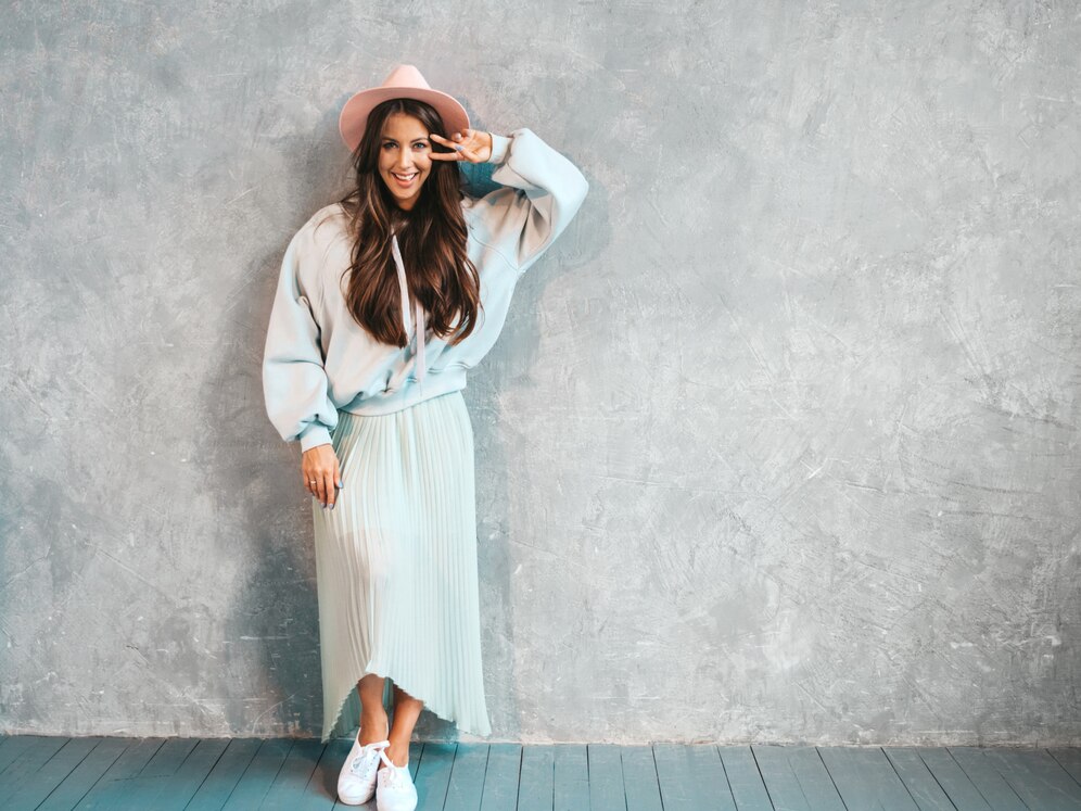 A woman poses against a textured gray wall, wearing a light blue oversized sweatshirt, a pleated skirt, and a pink hat.