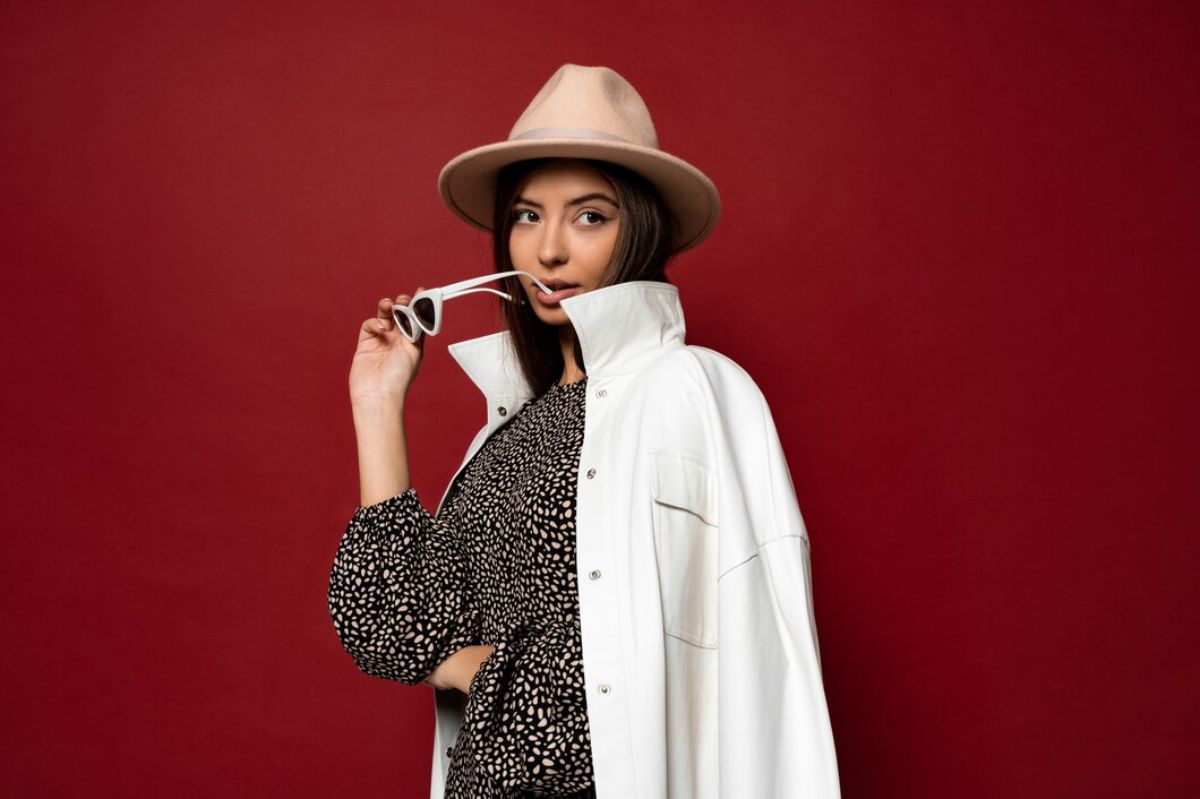 A woman poses confidently wearing a white jacket over a black spotted dress and holding stylish sunglasses.