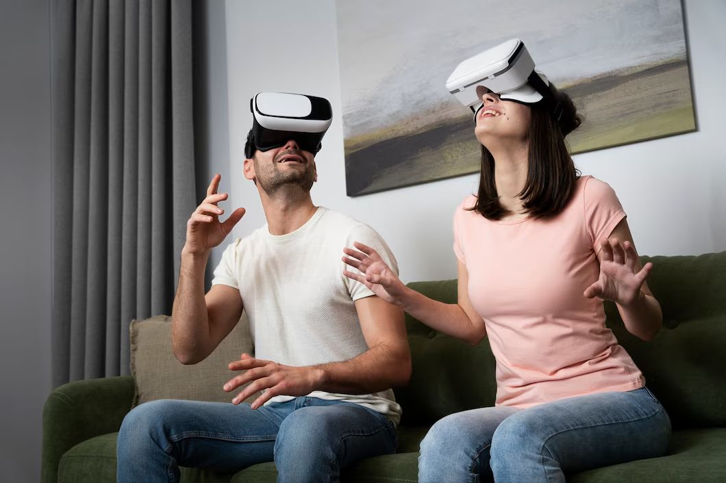  A man and a woman sitting on a green couch, both wearing virtual reality headsets, gesturing excitedly in a modern living room.