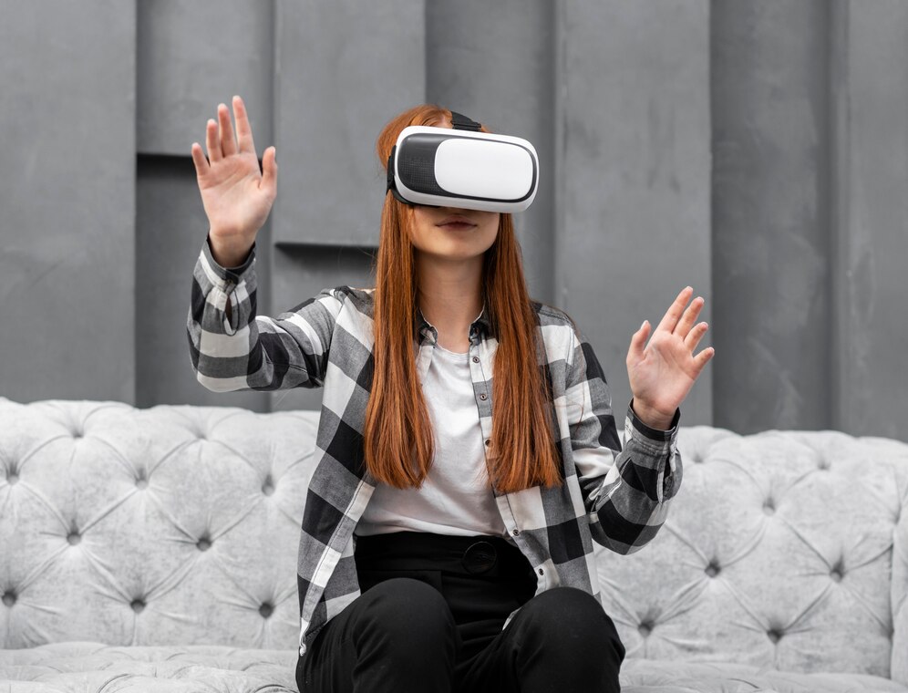 A woman with long reddish-brown hair wears a VR headset, gesturing with both hands while sitting on a plush gray sofa.