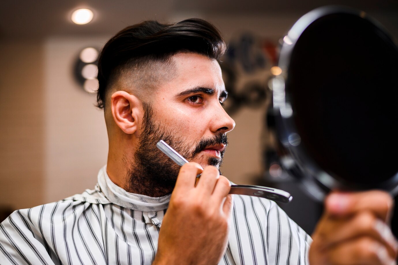 person in a striped barber cape holds a mirror, examining their hair
