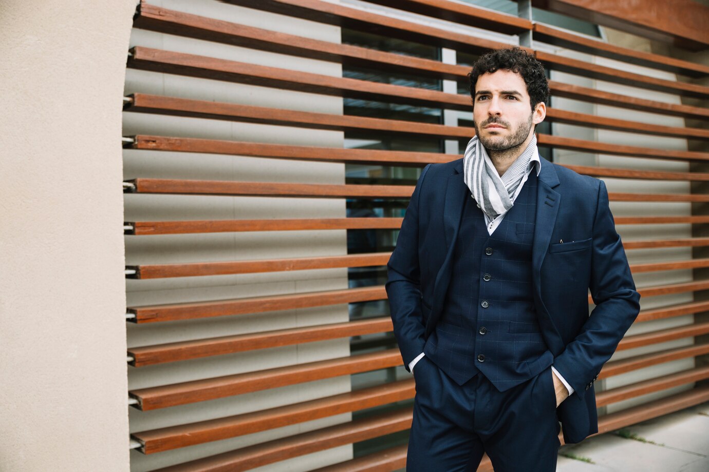 A well-dressed man in a tailored suit and scarf stands confidently against a backdrop of wooden slats.