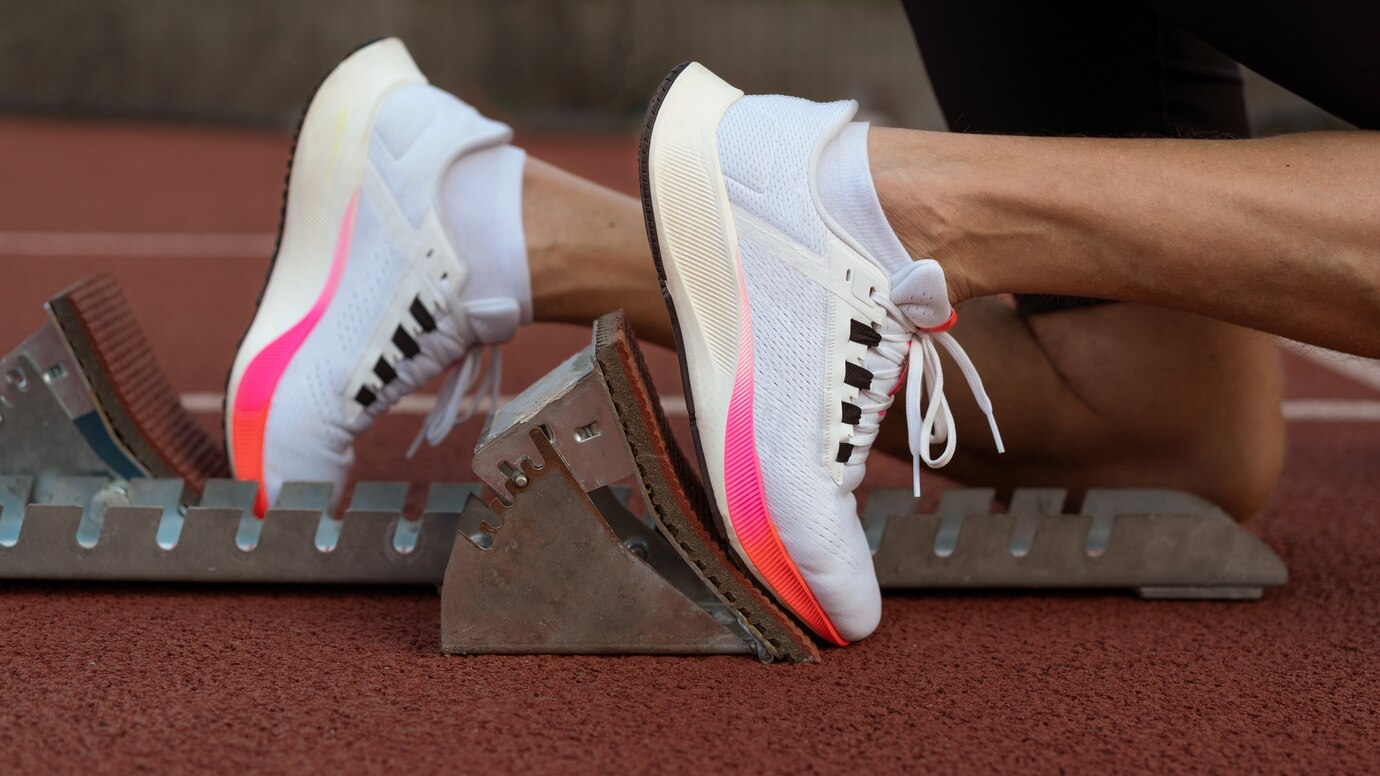 Close-up of a sprinter's foot in a sleek white and pink shoe, positioned in a starting block, ready for a race on a red track.