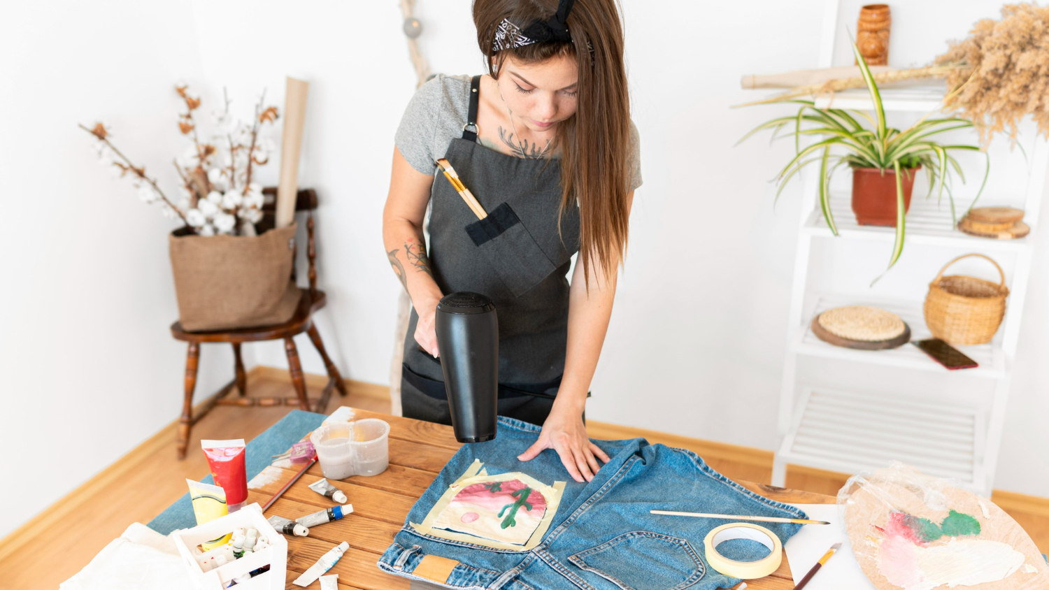 A person in an apron uses a hairdryer on decorated denim, surrounded by art supplies in a creative workspace.