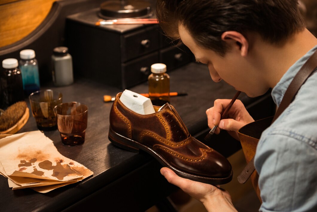 A shoemaker carefully paints a polished brown leather shoe at a workbench surrounded by various tools and materials.