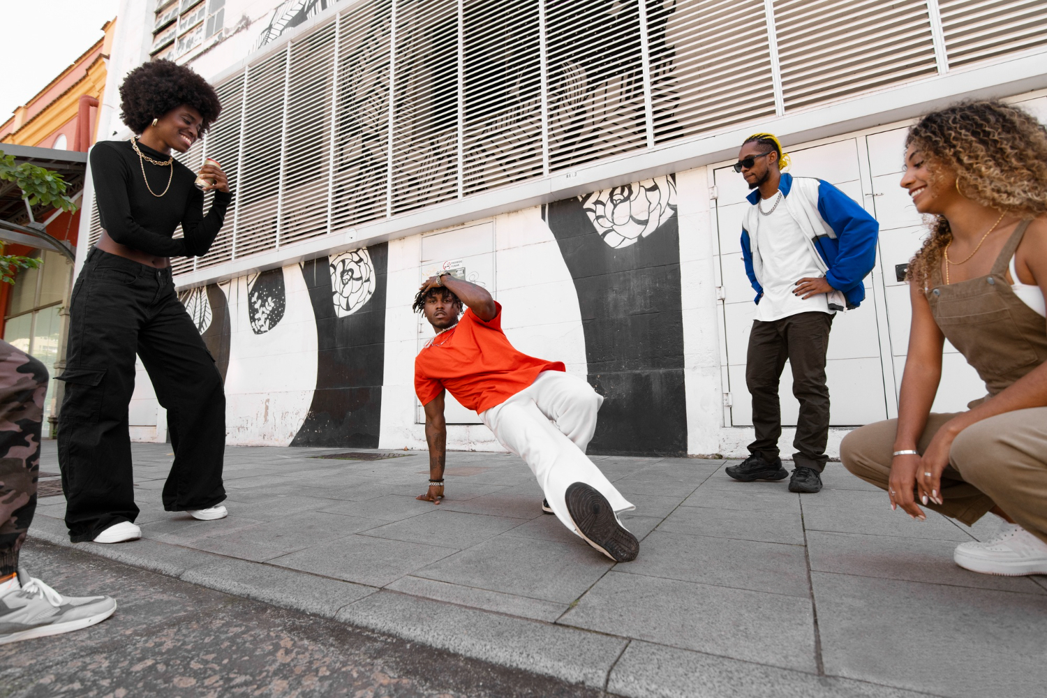 A group of friends watches and supports a dancer performing a breakdance move on a city sidewalk, with vibrant urban art in the background.