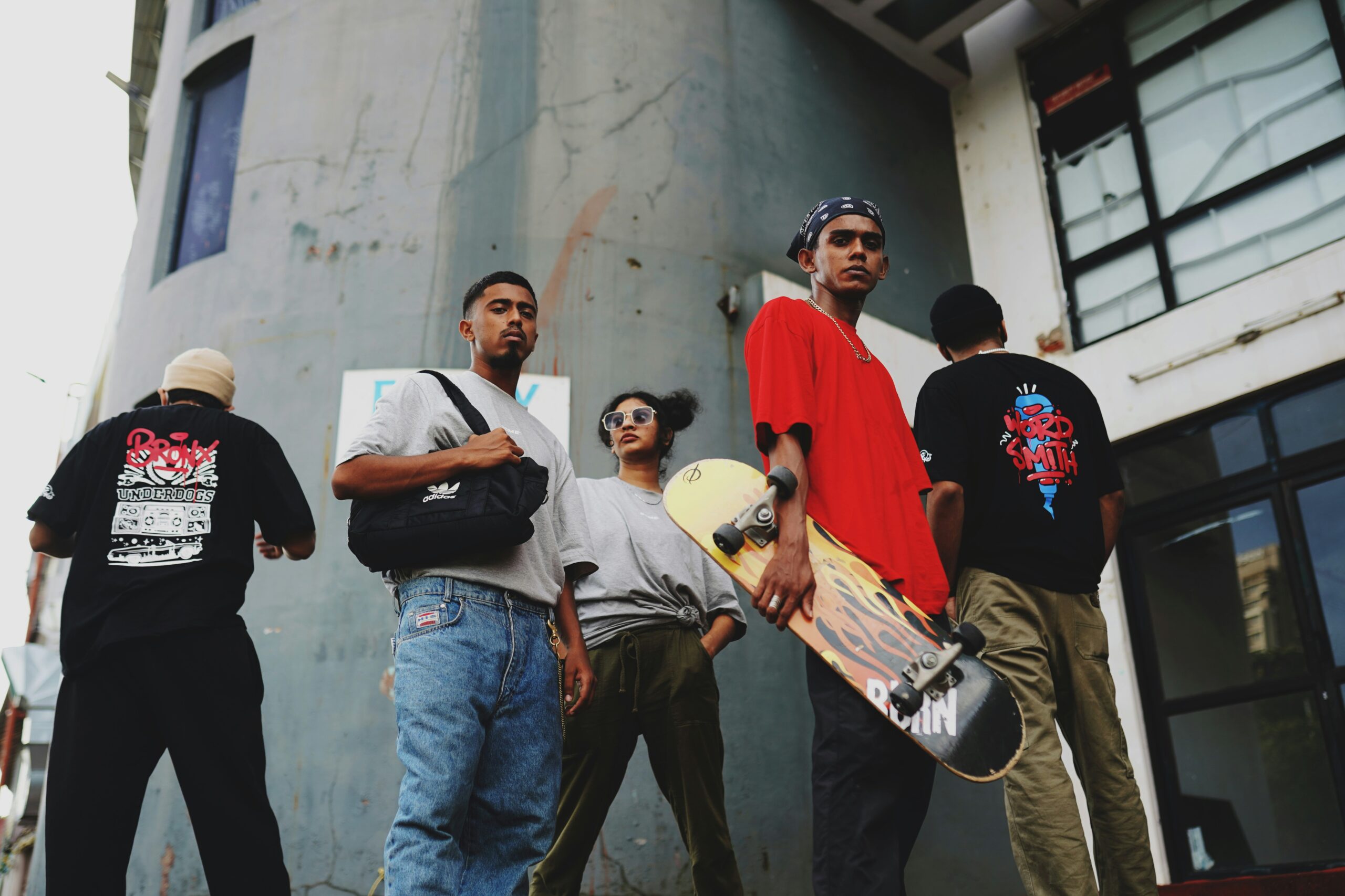 A group of six stylish individuals stand in front of a building, showcasing skateboards and urban fashion.
