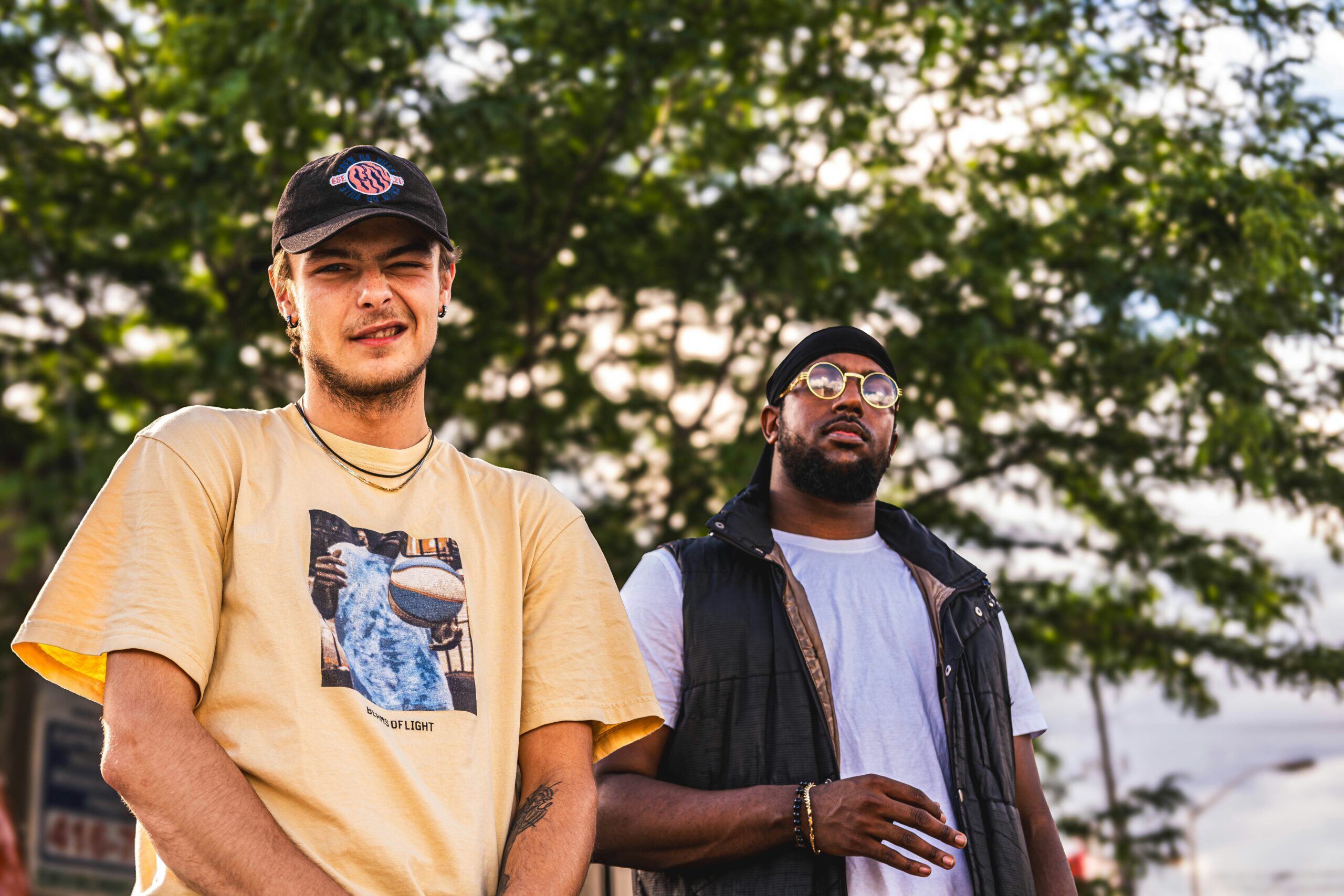 Two men stand outdoor greenery, one wearing t-shirt and the other in a white shirt.
