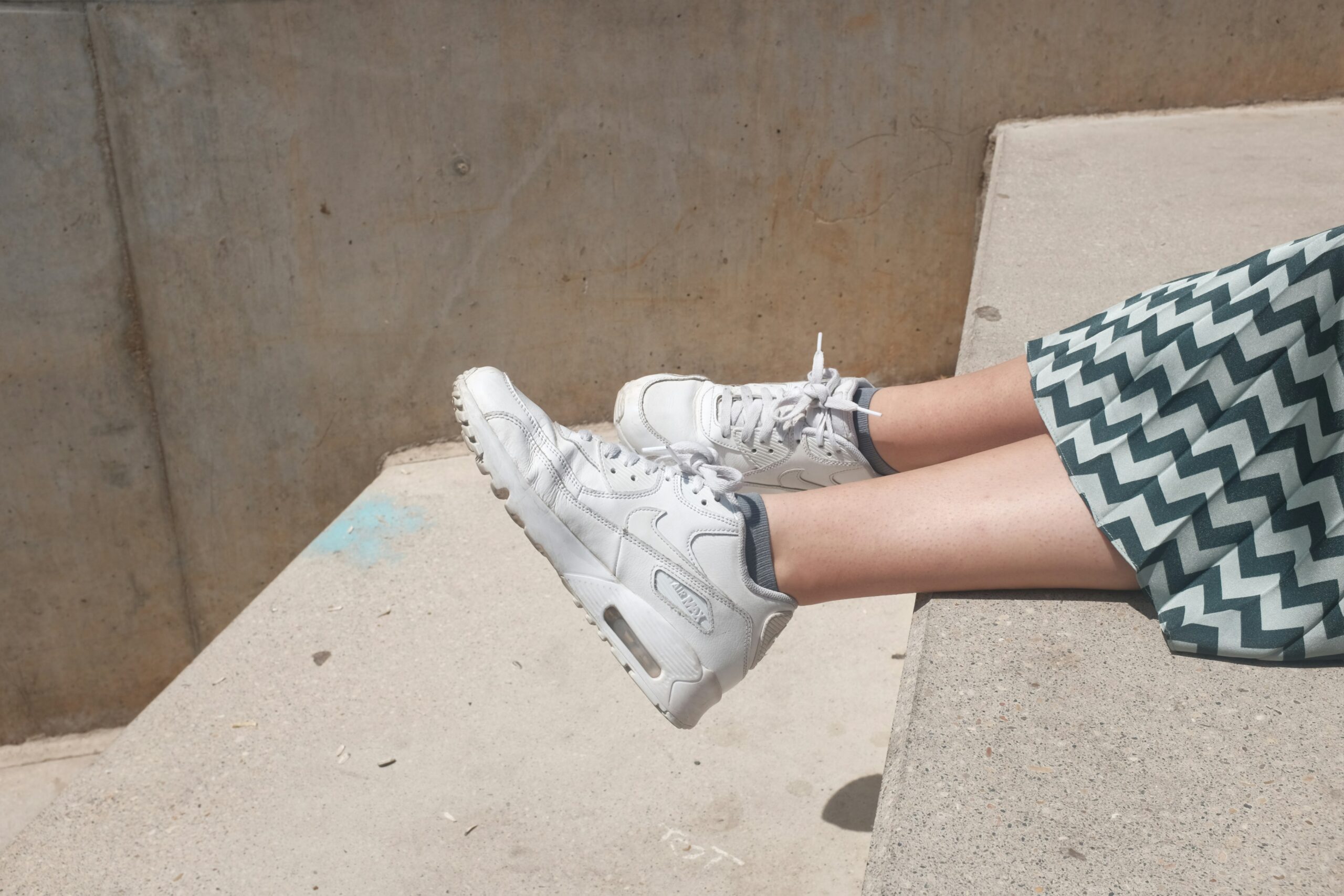 A pair of white Nike Air Max sneakers rests on a concrete ledge, with a zigzag patterned skirt visible in the frame.