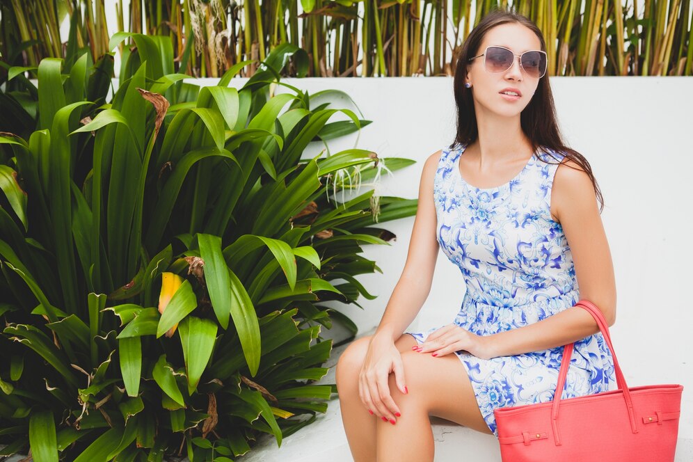 Woman in a blue floral dress and sunglasses sitting by lush green plants with a red tote bag.