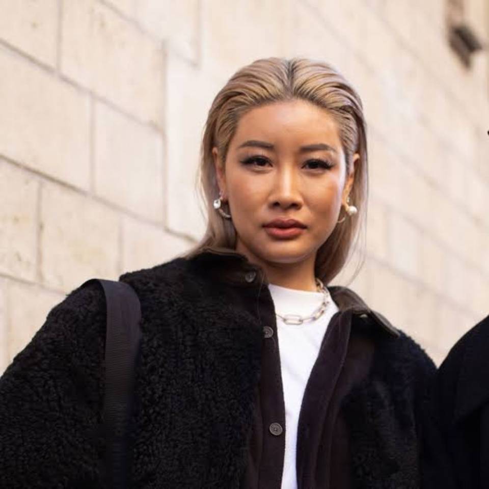 Young woman with styled hair in a chic black outfit, wearing earrings and a necklace, looking directly at the camera.