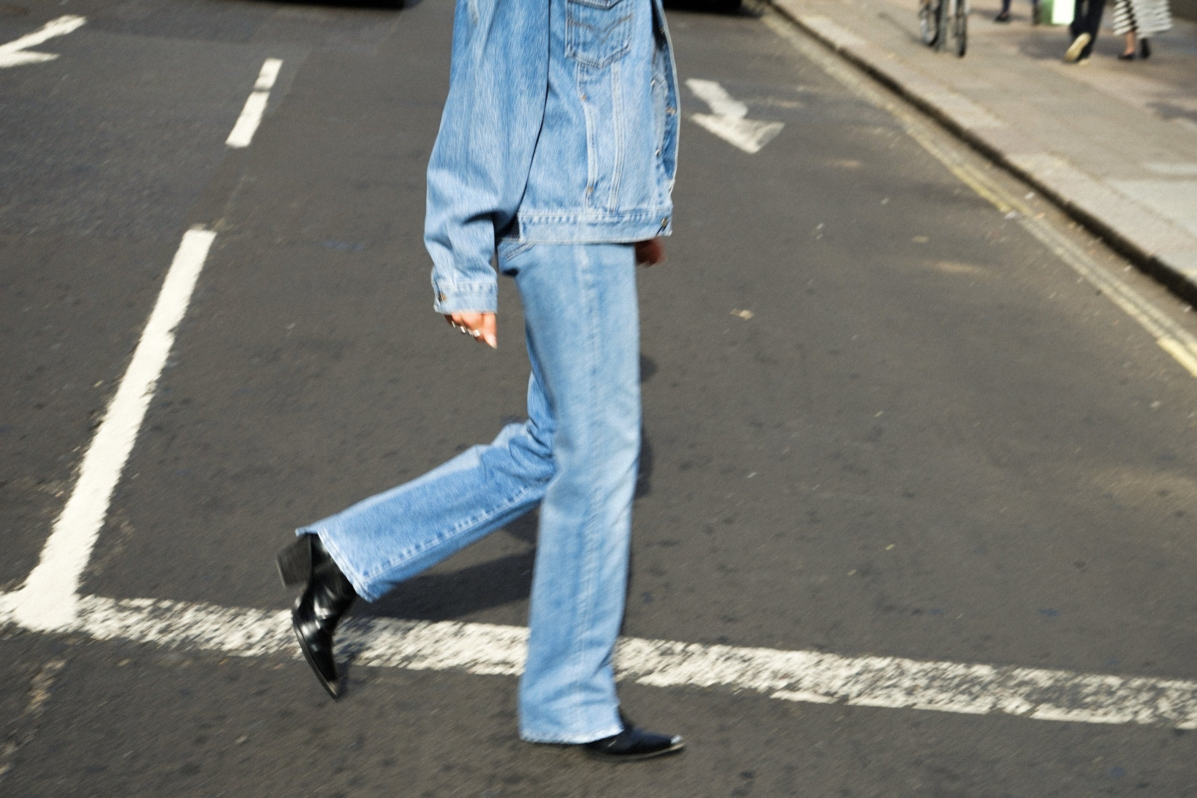 Person in denim jeans and jacket walking across a street, mid-step, on a sunny day.