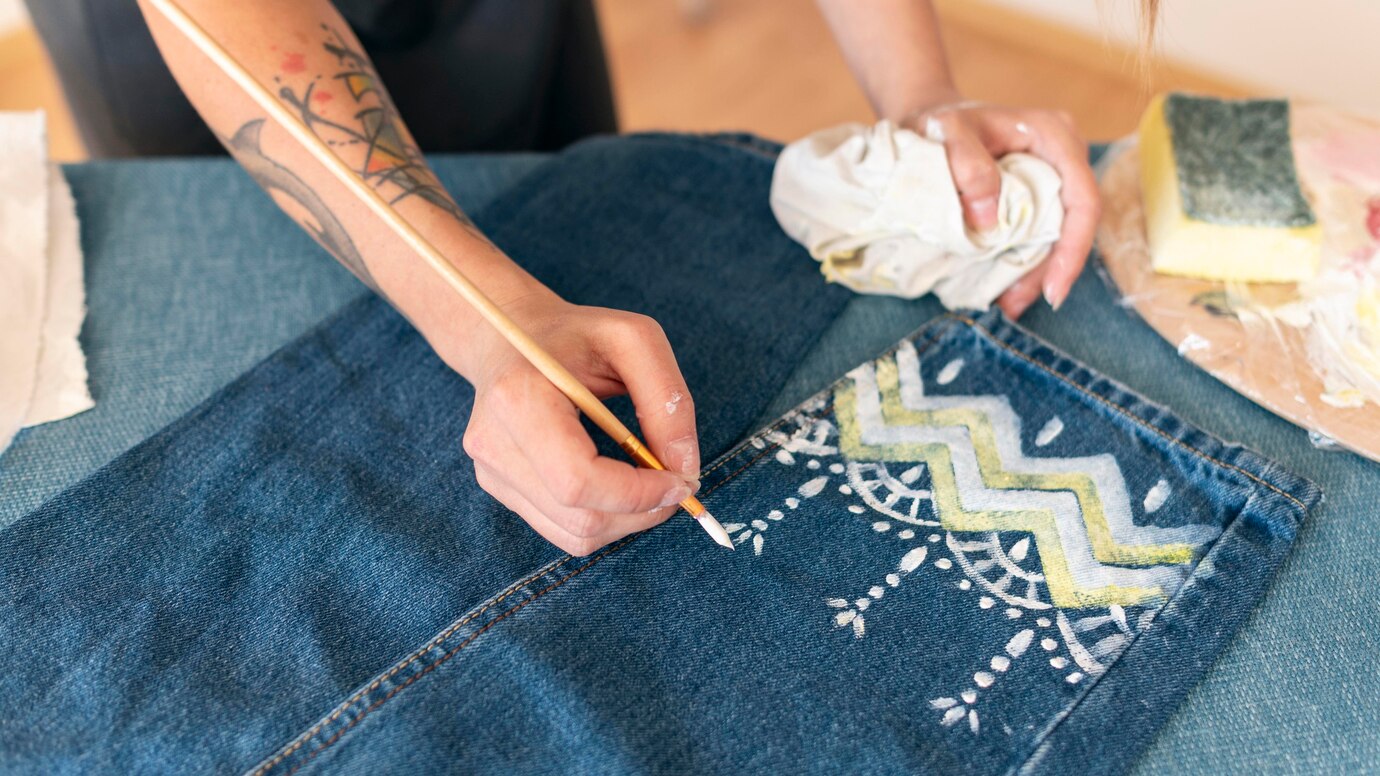 A person paints a patterned design in white and yellow on denim fabric, using a paintbrush, while holding a cloth in the other hand.