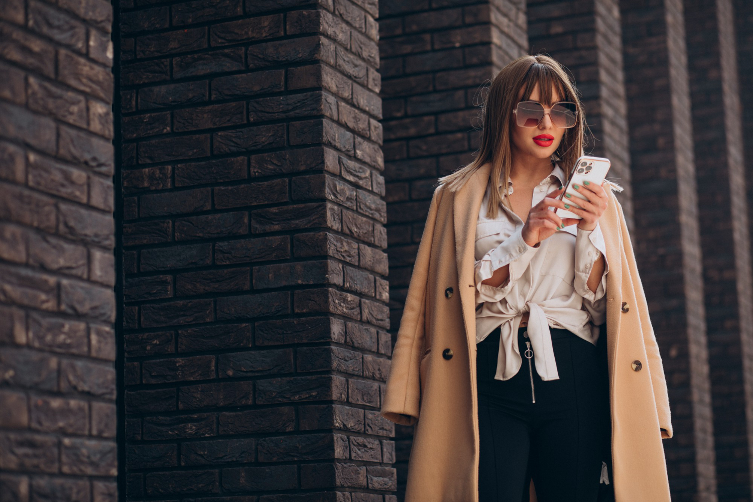 A stylish woman in sunglasses and a trench coat using a smartphone by a dark brick wall.