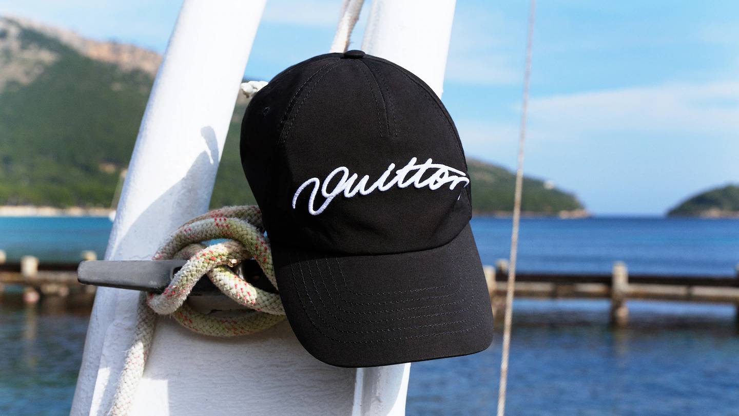 Black baseball cap with white script, hanging on a boat's white railing, with a serene sea and hills in the background.