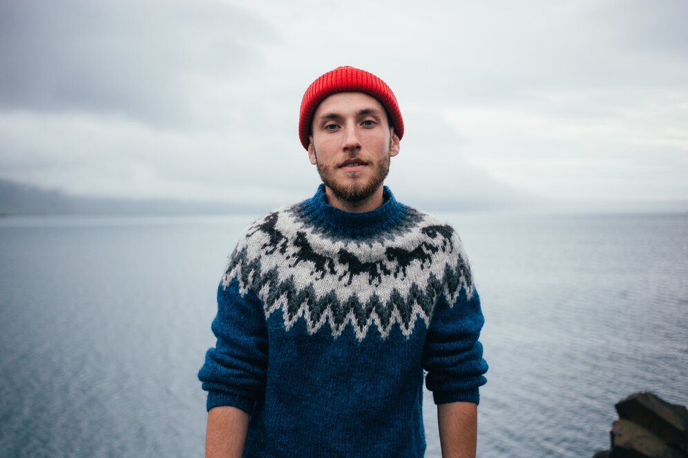 Young man in a red beanie and blue sweater standing by a foggy lake.