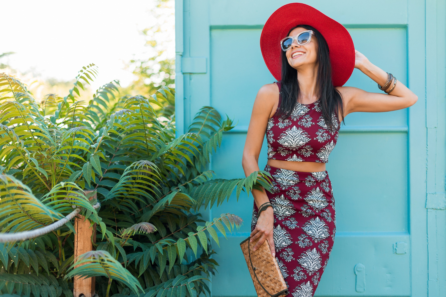 Woman in a red hat and sunglasses posing with a smile near tropical foliage and a pale blue door.