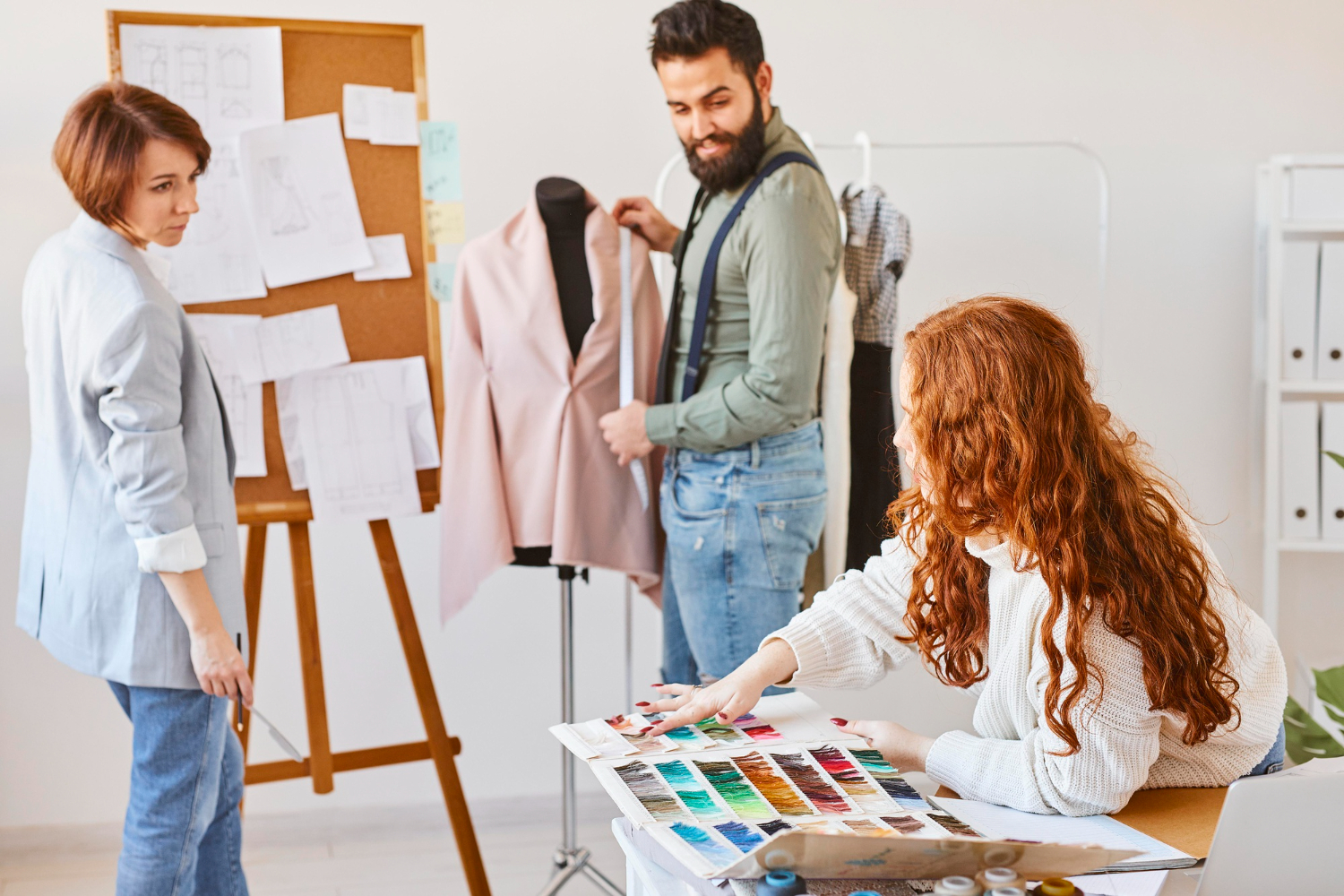 Three fashion designers discussing over fabric samples in a bright workshop with mannequin and clothes sketches in the background.