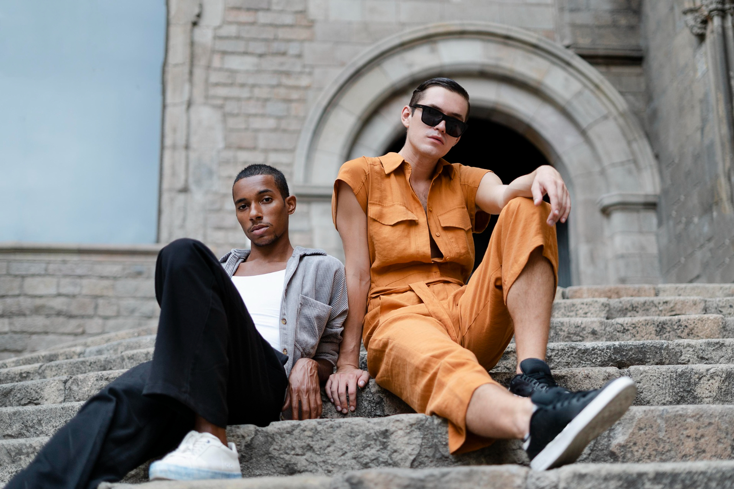 Two young men sitting casually on stone steps, one in orange outfit and sunglasses, the other in a gray jacket.